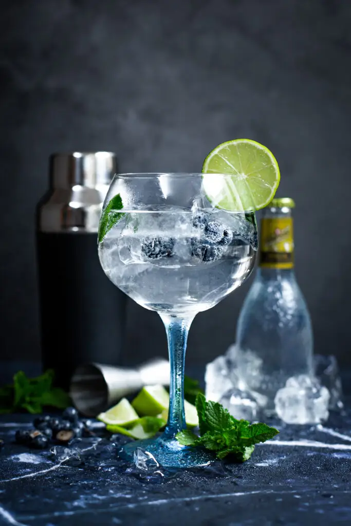 A glass of gin and tonic with mint, lime wedges and ice on a grey background